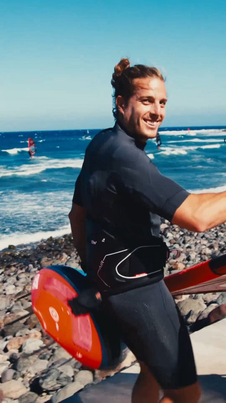 Federico is watching and smiling while holding a surfboard